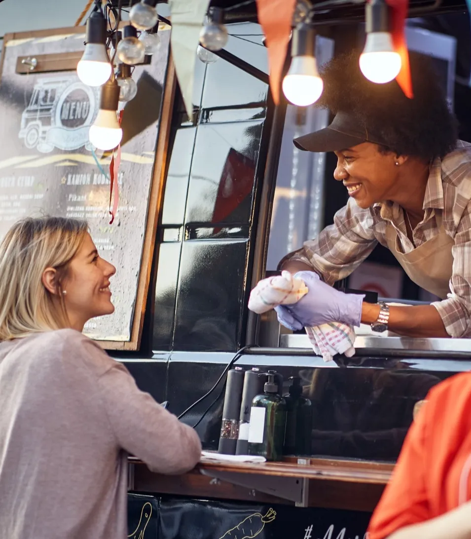 Eine lächelnde Foodtruck-Mitarbeiterin lehnt sich mit einem Geschirrtuch in der Hand aus dem Verkaufsfenster, umgeben von Lichterkette und Menütafel