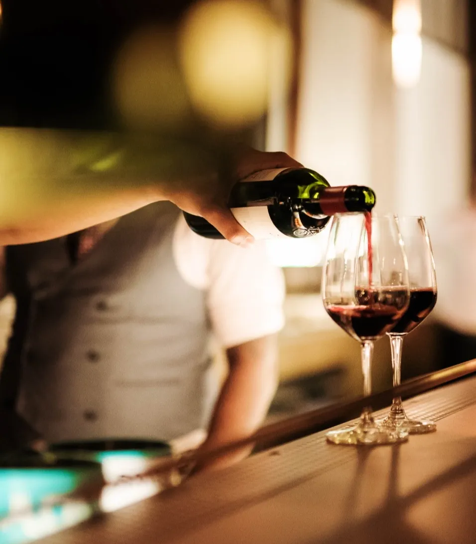 Eine Person in einer Weste schenkt Rotwein in zwei Gläser an einer schwach beleuchteten Bar ein.