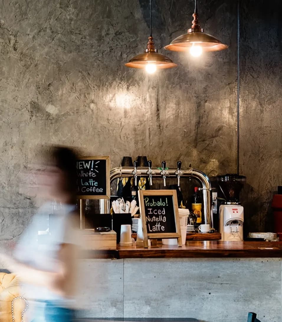 Unscharfe Person geht an einem gemütlichen Café-Tresen mit Kreidetafeln, industriellen Leuchten und rustikaler Betonwand vorbei.